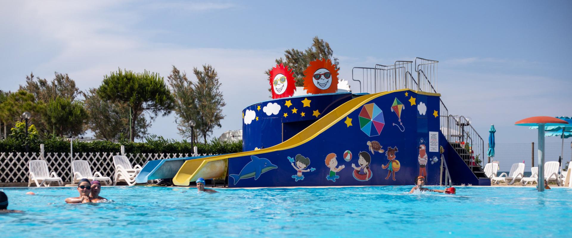 Piscina con scivolo colorato e decorazioni, bambini che nuotano e giocano.