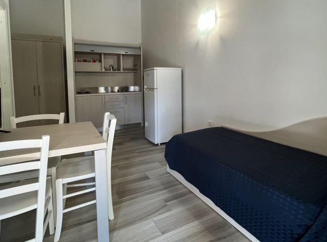 Monolocale con cucina, tavolo, letto singolo e pavimento in legno chiaro.
