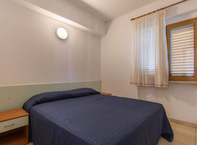 Camera semplice con letto matrimoniale, finestra e luce a parete.