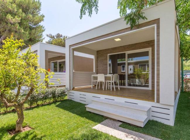 Moderna casa prefabbricata con veranda, circondata da un giardino verde e alberi.
