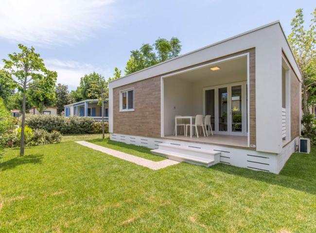 Moderna casa prefabbricata con veranda, circondata da giardino verde e alberi.