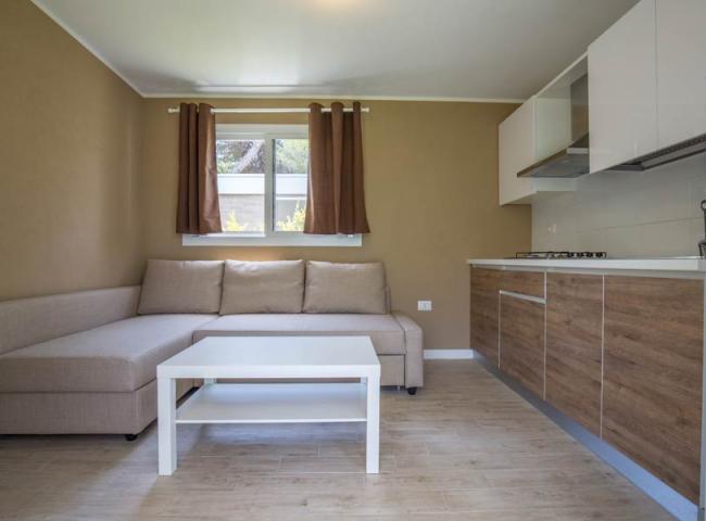 Soggiorno moderno con divano beige, tavolino bianco e cucina integrata in legno.