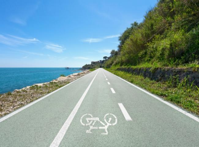 Pista ciclabile costiera con vista mare e vegetazione rigogliosa sotto un cielo azzurro.